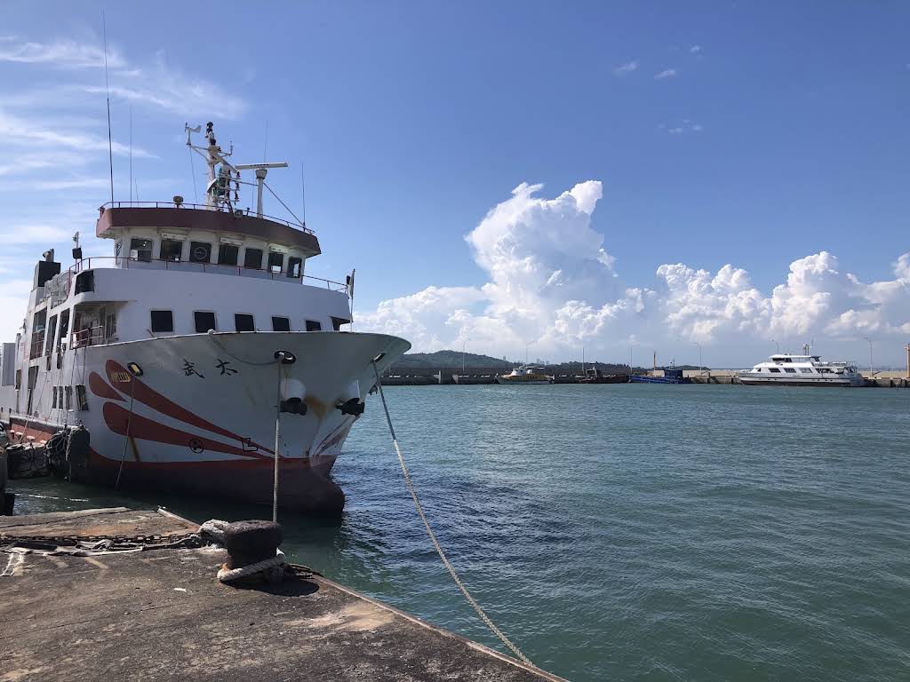 A white and red passenger ferry tied up at a Kinmen pier under a bright sky, the strait and the mainland shore beyond it